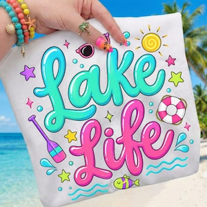 Puede incluir: Toalla de playa blanca con las palabras "Lake Life" en turquesa y rosa, rodeada de gráficos de dibujos animados de un sol, estrellas, un salvavidas y un pez. La toalla la sostiene una persona con pulseras coloridas y arte de uñas.