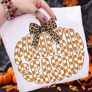 May include: A white t-shirt featuring a large graphic of a pumpkin shape filled with an orange and white checkered pattern. The pumpkin is topped with a leopard print bow. The text PUMPKIN SEASON is written in large, bold, orange and white letters.