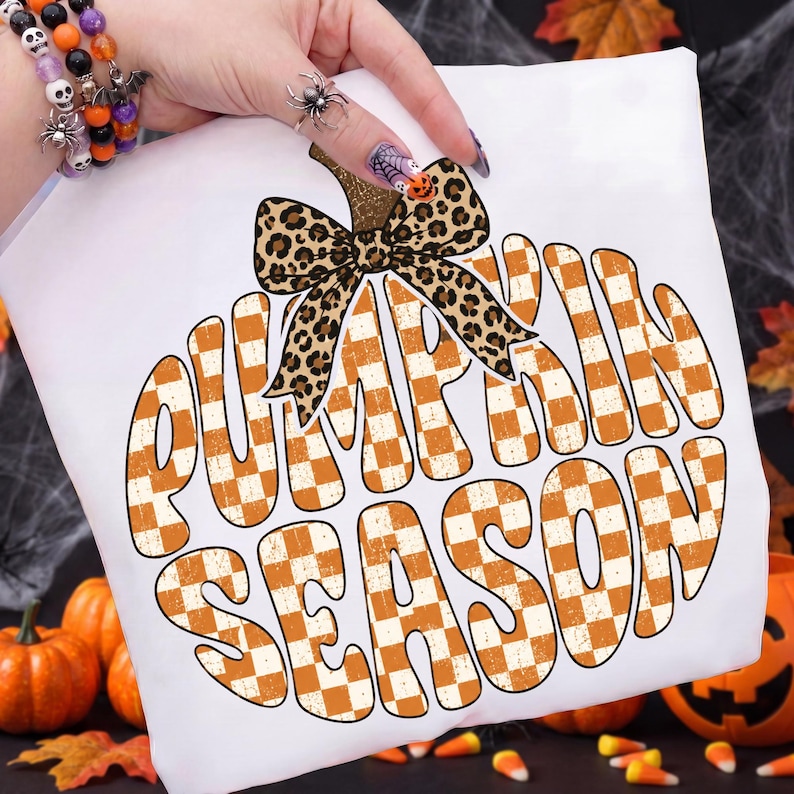 May include: A white t-shirt featuring a large graphic of a pumpkin shape filled with an orange and white checkered pattern. The pumpkin is topped with a leopard print bow. The text PUMPKIN SEASON is written in large, bold, orange and white letters.