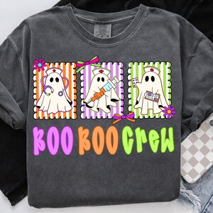 May include: Dark gray t-shirt with a colorful graphic of three ghost nurses. Each ghost is in a different frame with orange, purple, and green stripes. The text "BOO BOO CREW" is below the ghosts in pink, orange, and green.