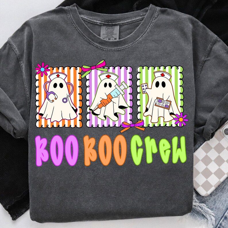 May include: Dark gray t-shirt with a colorful graphic of three ghost nurses. Each ghost is in a different frame with orange, purple, and green stripes. The text "BOO BOO CREW" is below the ghosts in pink, orange, and green.