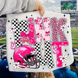 May include: A white graphic featuring the words "PINK NIT" in pink, black, and white. The design includes a pink football helmet, butterflies, flowers, and footballs. The background shows a football stadium.