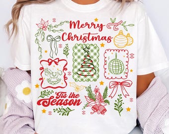 May include: White t-shirt with a festive Christmas design. The design includes the text "Merry Christmas" and "'Tis the Season" in red script, along with Christmas tree, ornaments, and bow graphics in green and red.
