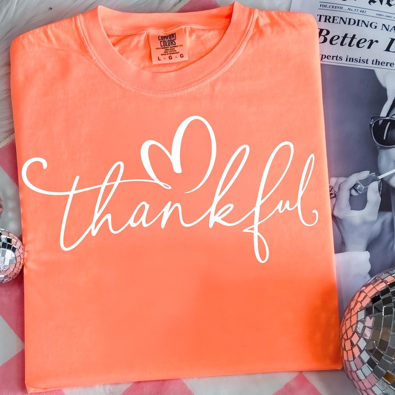 May include: Coral-colored t-shirt with the word "thankful" in white script, featuring a heart above the word. The shirt has a crew neck and short sleeves. The Comfort Colors brand label is visible.