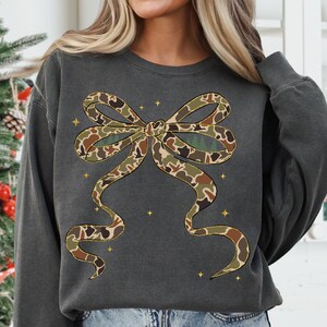 May include: Dark gray sweatshirt featuring a large bow design in a camouflage pattern. The bow has brown, green, and tan colors. Small gold stars are scattered around the bow. The sweatshirt is a casual style.