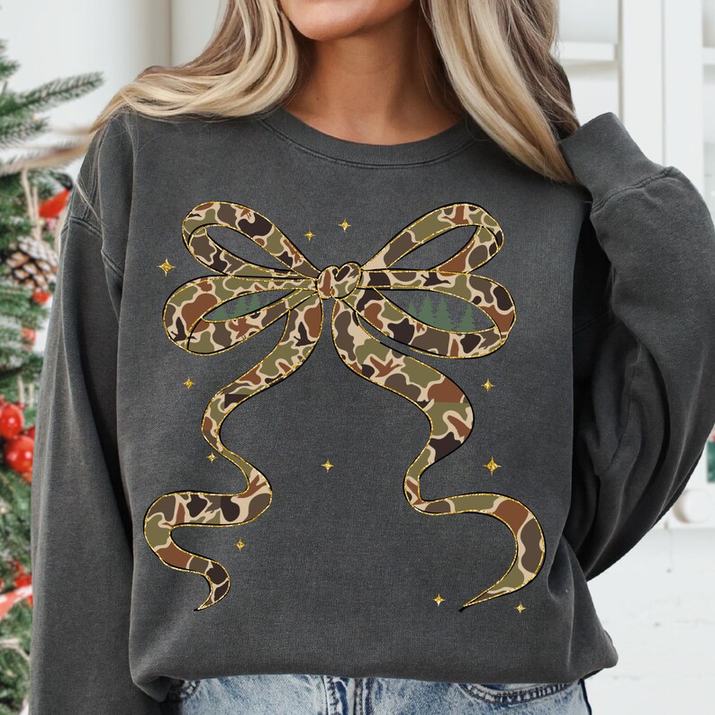 May include: Dark gray sweatshirt featuring a large bow design in a camouflage pattern. The bow has brown, green, and tan colors. Small gold stars are scattered around the bow. The sweatshirt is a casual style.