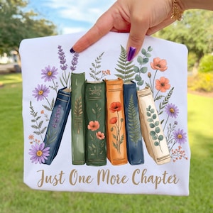 May include: White sweatshirt featuring a colorful illustration of books with floral accents. The design includes lavender, daisies, and poppies. The text "Just One More Chapter" is printed in a script font, perfect for book lovers.