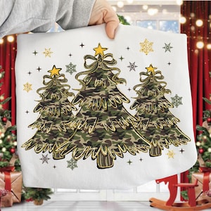 Può includere: Cuscino bianco con tre alberi di Natale stilizzati in un motivo mimetico. Ogni albero ha una stella e un fiocco dorati, circondati da fiocchi di neve e stelle dorate. Il cuscino è tenuto da una persona.
