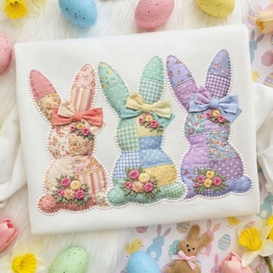 May include: A white fabric featuring three appliqued bunny figures. Each bunny is made of patchwork fabric in pastel colors, with a bow and floral embellishments. The bunnies are outlined with a pearl-like trim, creating a charming Easter theme.