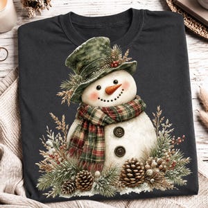 May include: A dark gray t-shirt featuring a graphic of a snowman wearing a green top hat and a red and green plaid scarf. The snowman has a carrot nose, two dark buttons on its torso, and is surrounded by pine branches, berries, and pinecones.