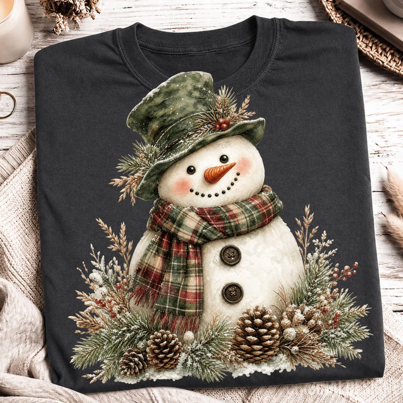 May include: A dark gray t-shirt featuring a graphic of a snowman wearing a green top hat and a red and green plaid scarf. The snowman has a carrot nose, two dark buttons on its torso, and is surrounded by pine branches, berries, and pinecones.