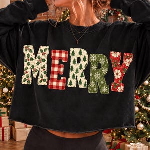 May include: A black long-sleeve sweatshirt featuring the word MERRY in large block letters across the chest. Each letter has a distinct holiday pattern: holly leaves, red and white buffalo plaid, green pine trees, white snowflakes, and red poinsettias.