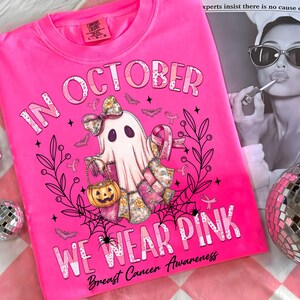 May include: Bright pink t-shirt with the text "IN OCTOBER WE WEAR PINK" and "Breast Cancer Awareness". The shirt features a ghost with a pumpkin bucket, bats, and pink ribbons. The shirt is a Comfort Colors brand.