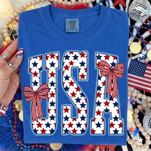 May include: Blue t-shirt with the word "USA" in white letters filled with red and blue stars. Red, white, and blue bows are on the letters. The shirt has a crew neck and a tag that says "Comfort Colors".