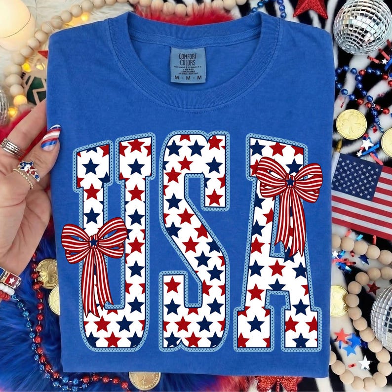 May include: Blue t-shirt with the word "USA" in white letters filled with red and blue stars. Red, white, and blue bows are on the letters. The shirt has a crew neck and a tag that says "Comfort Colors".