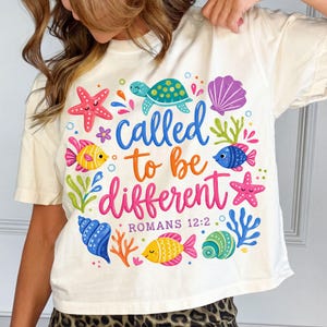 May include: Cream-colored t-shirt with a colorful aquatic design. The shirt features a turtle, starfish, fish, and seashells, along with the words "called to be different" and "Romans 12:2".