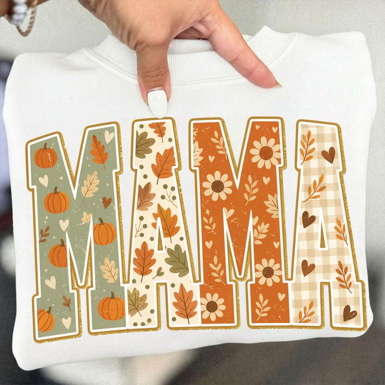 May include: A white crewneck sweatshirt featuring the word MAMA in large block letters. Each letter has a unique fall-themed pattern including orange pumpkins on sage green, autumn leaves, sunflowers on burnt orange, and beige buffalo plaid.