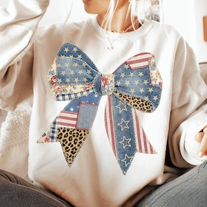 May include: White sweatshirt featuring a large bow design. The bow is a patchwork of denim, floral, leopard print, and patriotic patterns with stars and stripes. The bow is the focal point of the sweatshirt.