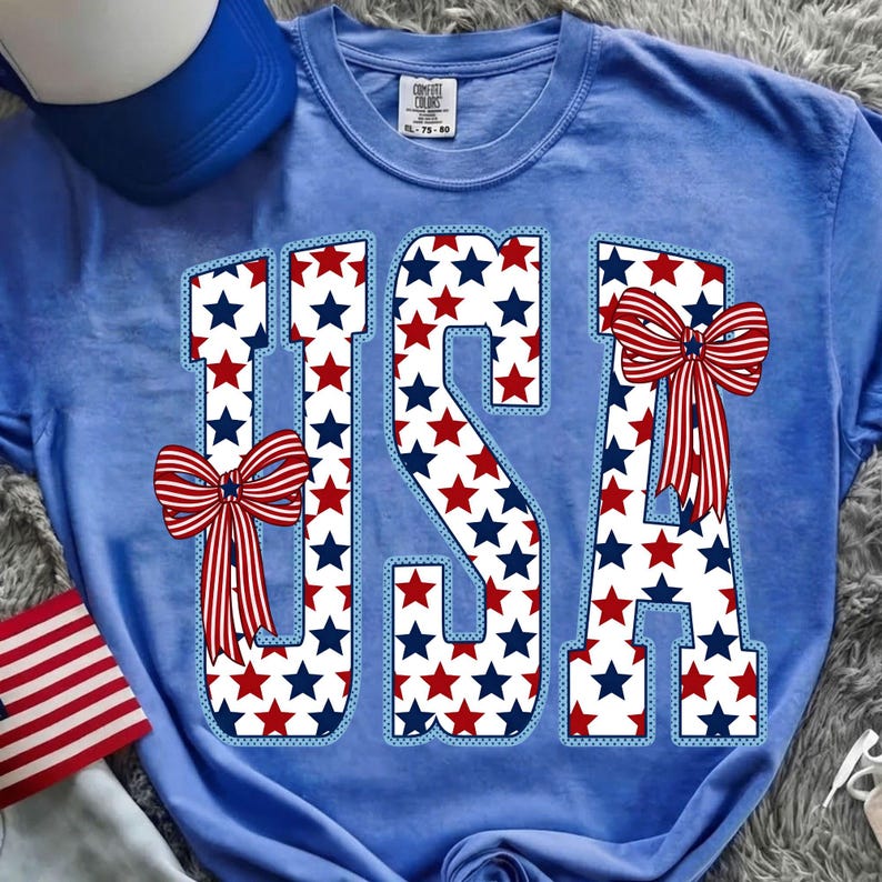 May include: Light blue t-shirt with the word "USA" in a star-filled design. The letters are outlined in light blue and have red, white, and blue stars. Red, white, and blue bows are on the letters. A blue hat and a small American flag are in the background.