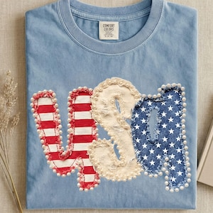 May include: Light blue t-shirt with the word "USA" in red and white stripes, cream, and blue denim with white stars. The letters are outlined with small white beads. The shirt has a crew neck and a small tag that says "Comfort Colors".