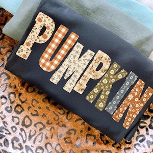 May include: A dark gray t-shirt featuring the word PUMPKIN printed in large letters. Each letter has a unique pattern, including orange pumpkins, brown and white buffalo plaid, and various floral designs in shades of orange, green, and white.