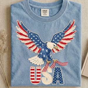 Op de afbeelding: Lichtblauw T-shirt met een geborduurde adelaar met uitgespreide vleugels, met de sterren en strepen van de Amerikaanse vlag. Het woord "USA" is ook in rood en wit geborduurd. Het shirt heeft een ronde hals en een label met "Comfort Colors".