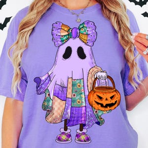 Op de afbeelding: Een paars t-shirt met een grillig spookontwerp. Het spook is versierd met patchworkdetails, een grote strik en houdt een pompoenemmer vast. Het ontwerp bevat verschillende patronen en kleuren, perfect voor Halloween.
