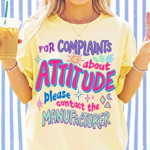 Puede incluir: Camiseta amarilla clara con el texto "FOR COMPLAINTS ABOUT ATTITUDE PLEASE CONTACT THE MANUFACTURER" en varios colores. El dise&ntilde;o incluye un sol sonriente, estrellas y otros elementos decorativos.