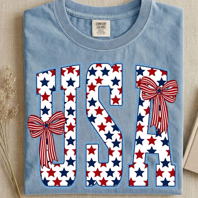 May include: Light blue t-shirt with the word "USA" in a patriotic design. The letters are filled with red and blue stars and outlined in blue. Red, white, and blue bows are attached to the letters.
