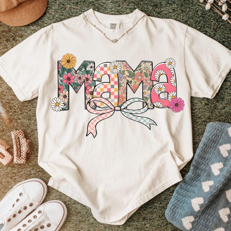 May include: White t-shirt with the word "Mama" in a floral design. The letters are filled with flowers and a checkered pattern. A pink and blue bow is below the word. The shirt has a crew neck and short sleeves.
