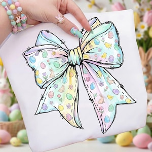 May include: White t-shirt with a large, pastel-coloured bow design. The bow is decorated with Easter eggs, bunnies, chicks, and flowers. The design has a hand-drawn aesthetic with black outlines and stitched details, ideal for Easter.