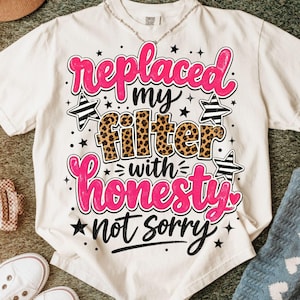 May include: A white short-sleeve t-shirt featuring the text "replaced my filter with honesty not sorry" in pink, black, and leopard print lettering. The design includes black and white striped stars and small black stars scattered throughout.