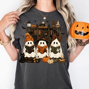 May include: A charcoal gray t-shirt featuring a graphic of three ghosts reading books in front of a bookshelf. The ghosts wear a beanie, a witch hat, and a bow. The design includes a black cat, pumpkins, candles, and books labeled Spells and Curses & Hexes.