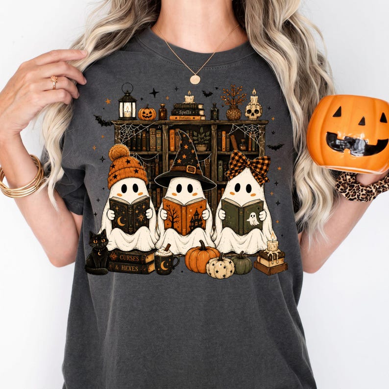 May include: A charcoal gray t-shirt featuring a graphic of three ghosts reading books in front of a bookshelf. The ghosts wear a beanie, a witch hat, and a bow. The design includes a black cat, pumpkins, candles, and books labeled Spells and Curses & Hexes.