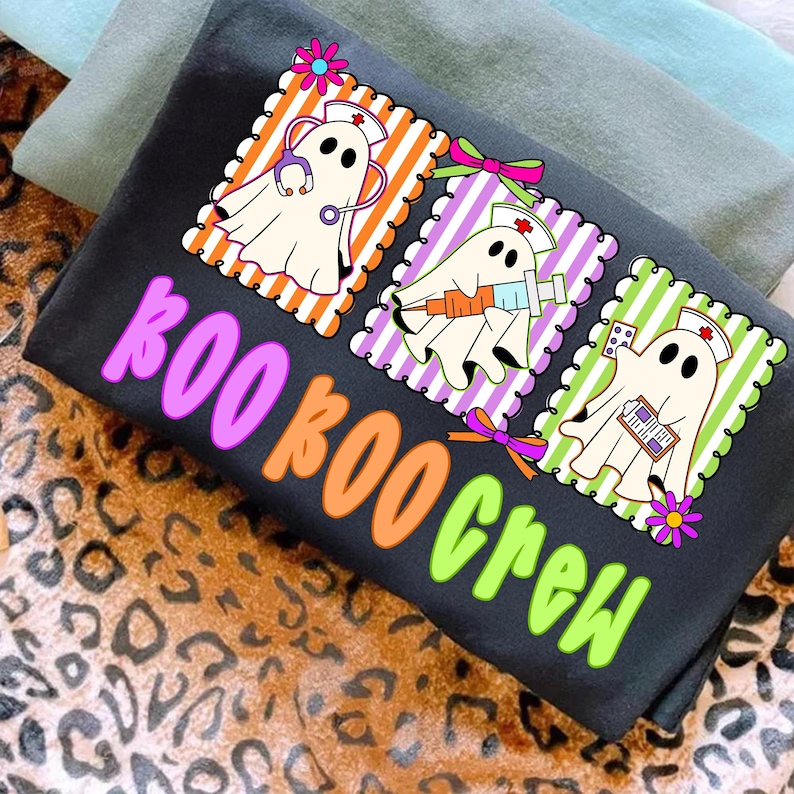 May include: Black t-shirt with a colorful graphic of three cartoon ghosts dressed as nurses, each holding medical tools. The text "BOO BOO CREW" is in bright pink, orange, and green. The shirt is folded on a leopard print surface.