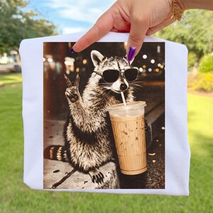 May include: White t-shirt with a sepia-toned image of a raccoon in sunglasses, holding an iced coffee and giving a peace sign. The background shows blurred city lights.