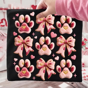 May include: Black square pillow featuring embroidered pink paw prints with heart-shaped centers and bows. The design includes small pink hearts and cross accents, creating a charming, pet-themed aesthetic.