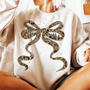 May include: Cream-colored sweatshirt featuring a large bow design in a camouflage pattern with a forest scene in the center. The bow has brown, green, and tan tones. Small gold stars are scattered around the bow.