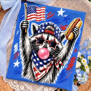 Puede incluir: Una impresión rectangular que muestra un mapache con una gorra de béisbol estrellada, gafas de sol en forma de estrella y un pañuelo con patrón de bandera. El mapache sostiene una bandera estadounidense y un perrito caliente, soplando una burbuja.