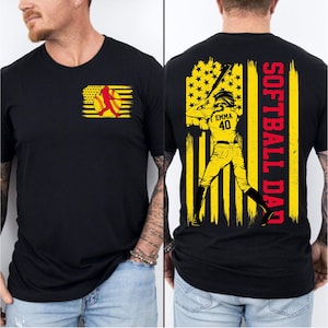 Op de afbeelding: Zwart T-shirt met een geel en rood softbalontwerp op de voorkant. De achterkant heeft een gele en rode afbeelding van een softbalspeler met de tekst "SOFTBALL DAD" in het rood.