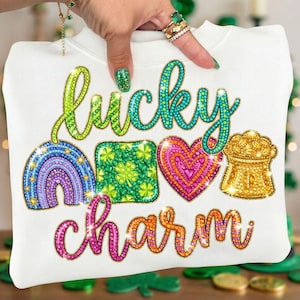 Puede incluir: Sudadera blanca con las palabras "lucky charm" en una escritura colorida y brillante. El diseño incluye un arcoíris, un trébol, un corazón y una olla de oro, todos con un efecto brillante. La sudadera es sostenida por una mano.