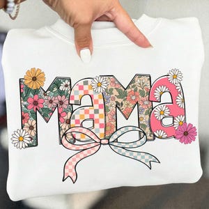 May include: White sweatshirt with the word "MAMA" in a floral design. The letters are filled with various floral patterns, a checkered pattern, and daisies. A pink and blue checkered bow is at the bottom of the letters.