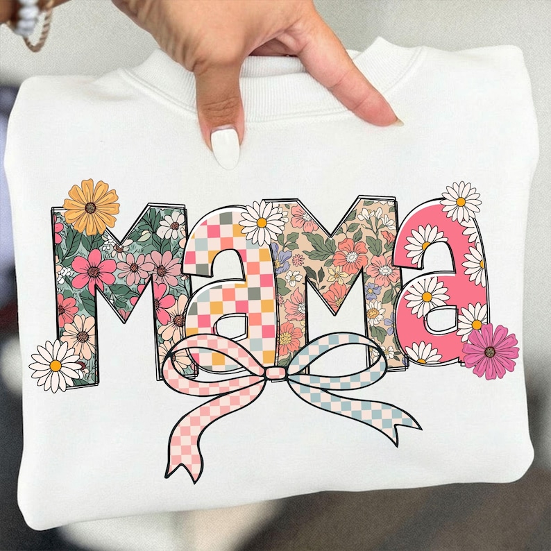 May include: White sweatshirt with the word "MAMA" in a floral design. The letters are filled with various floral patterns, a checkered pattern, and daisies. A pink and blue checkered bow is at the bottom of the letters.