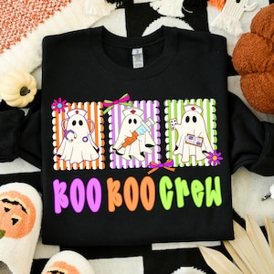 May include: Black sweatshirt with a colorful graphic featuring three ghost characters dressed as nurses, each with medical tools. The text "BOO BOO CREW" is below the graphic in pink, orange, and green. A fun Halloween design.