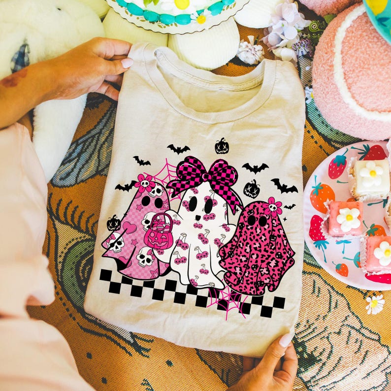 May include: A cream-colored t-shirt featuring three cartoon ghosts with pink accents. The ghosts have various patterns, including skulls, cherries, and leopard print. Black bats, spiderwebs, and pumpkins decorate the shirt, with a checkered border at the bottom. A pink bow adorns the central ghost.