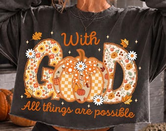 May include: A charcoal gray long-sleeve cropped sweatshirt featuring the text "With GOD All things are possible" in orange. The graphic includes a central pumpkin with a bow and floral patterns, surrounded by small white stars and autumn leaves.
