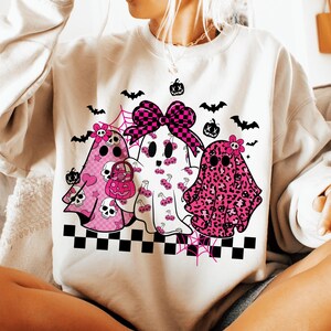 May include: A white sweatshirt featuring three ghost characters with pink accents. The ghosts have various patterns, including skulls, cherries, and leopard print. Black bats, pumpkins, and a checkered bow complete the Halloween-themed design.