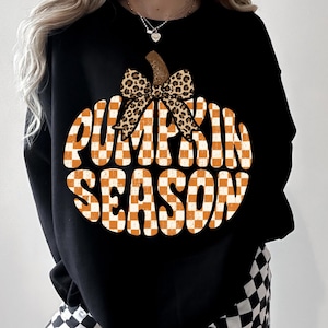 May include: A black long-sleeve sweatshirt featuring a large graphic of a pumpkin shape filled with an orange and white checkered pattern. The text PUMPKIN SEASON is printed in white across the pumpkin, topped with a leopard print bow.