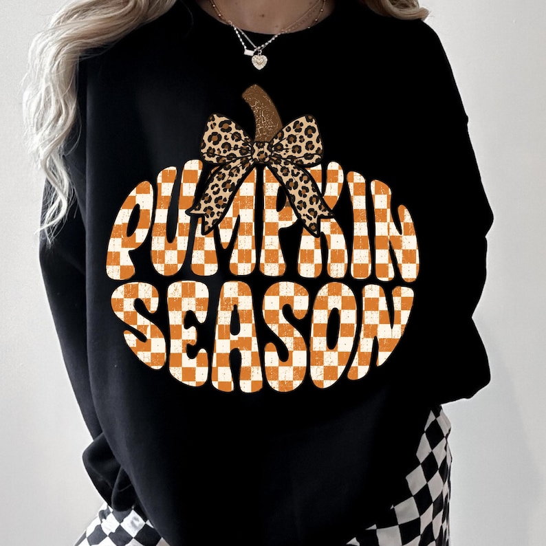 May include: A black long-sleeve sweatshirt featuring a large graphic of a pumpkin shape filled with an orange and white checkered pattern. The text PUMPKIN SEASON is printed in white across the pumpkin, topped with a leopard print bow.
