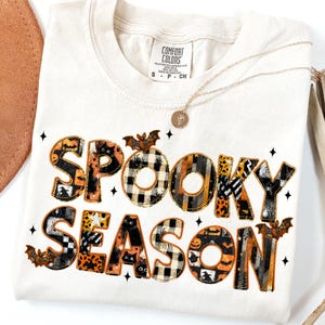 Puede incluir: Camiseta color crema con las palabras "SPOOKY SEASON" en un diseño de patchwork con murciélagos, arañas y calabazas. La camiseta tiene cuello redondo y mangas cortas. Un collar dorado con un colgante está sobre la camiseta.